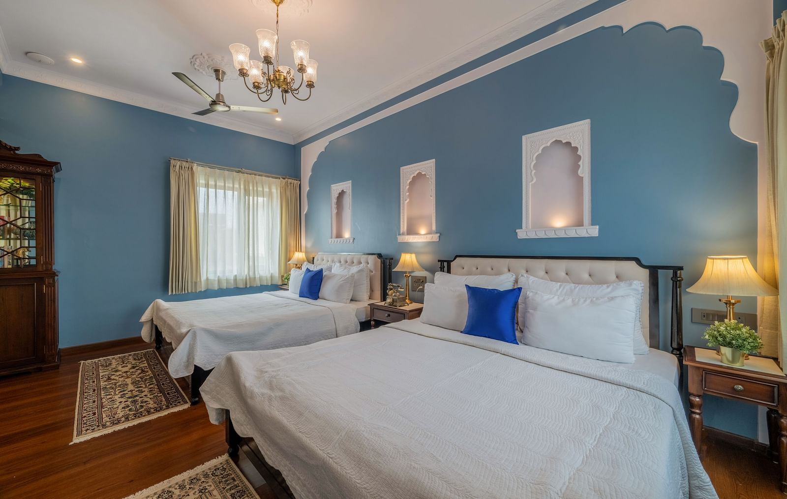 A spacious guest room at Dera Rawatsar, Jaipur, features two comfortable beds, elegant wall decor, wooden flooring, and a classic chandelier.