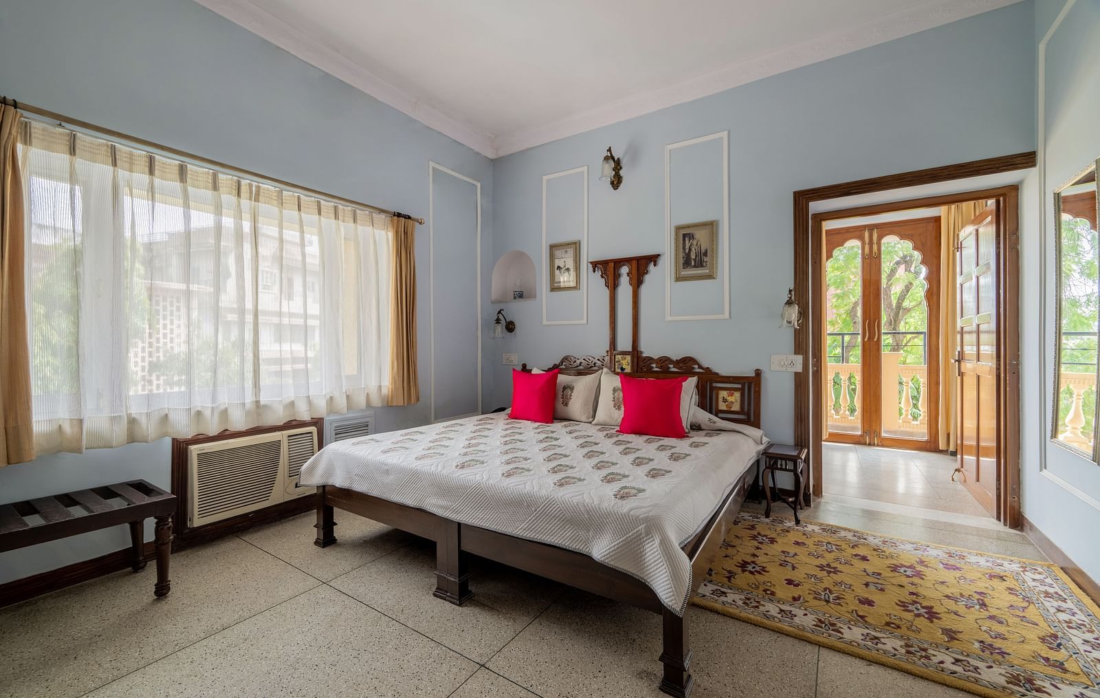 A bright and airy bedroom at Dera Rawatsar, Jaipur, features a comfortable bed, large windows with sheer curtains, and an ornate doorway leading outside.