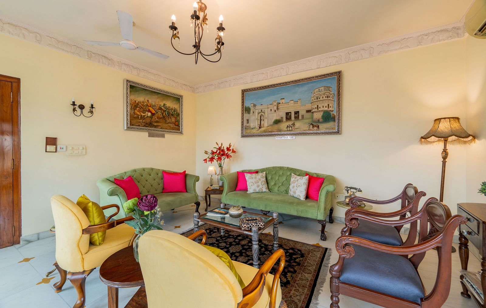 A living room with green and yellow sofas, colourful cushions, framed artwork, a chandelier, and a dark rug | Dera Rawatsar, Jaipur