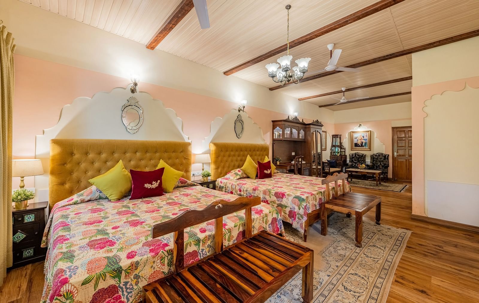 A grand suite at Dera Rawatsar, Jaipur, features two comfortable beds, a seating area with plush sofas, wooden beams, and elegant chandeliers.