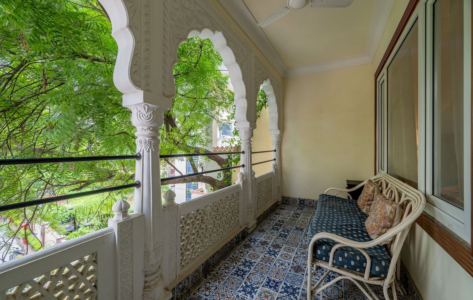 A charming balcony at Dera Rawatsar, Jaipur, features ornate arched railings, patterned tile flooring, and a comfortable cushioned bench with pillows.