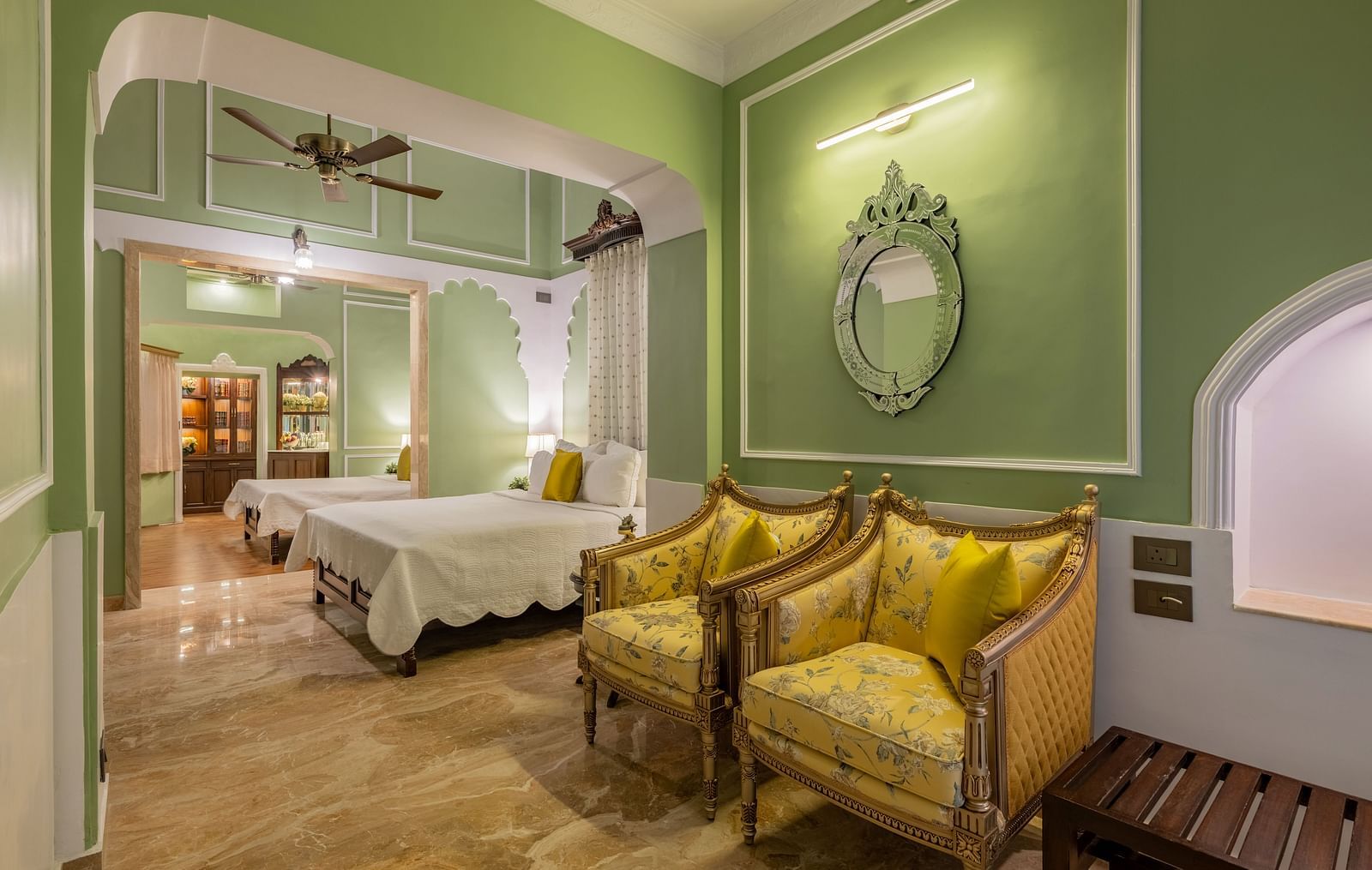 A charming suite at Dera Rawatsar, Jaipur, features two comfortable beds, stylish armchairs, an ornate mirror, and elegant archway details.