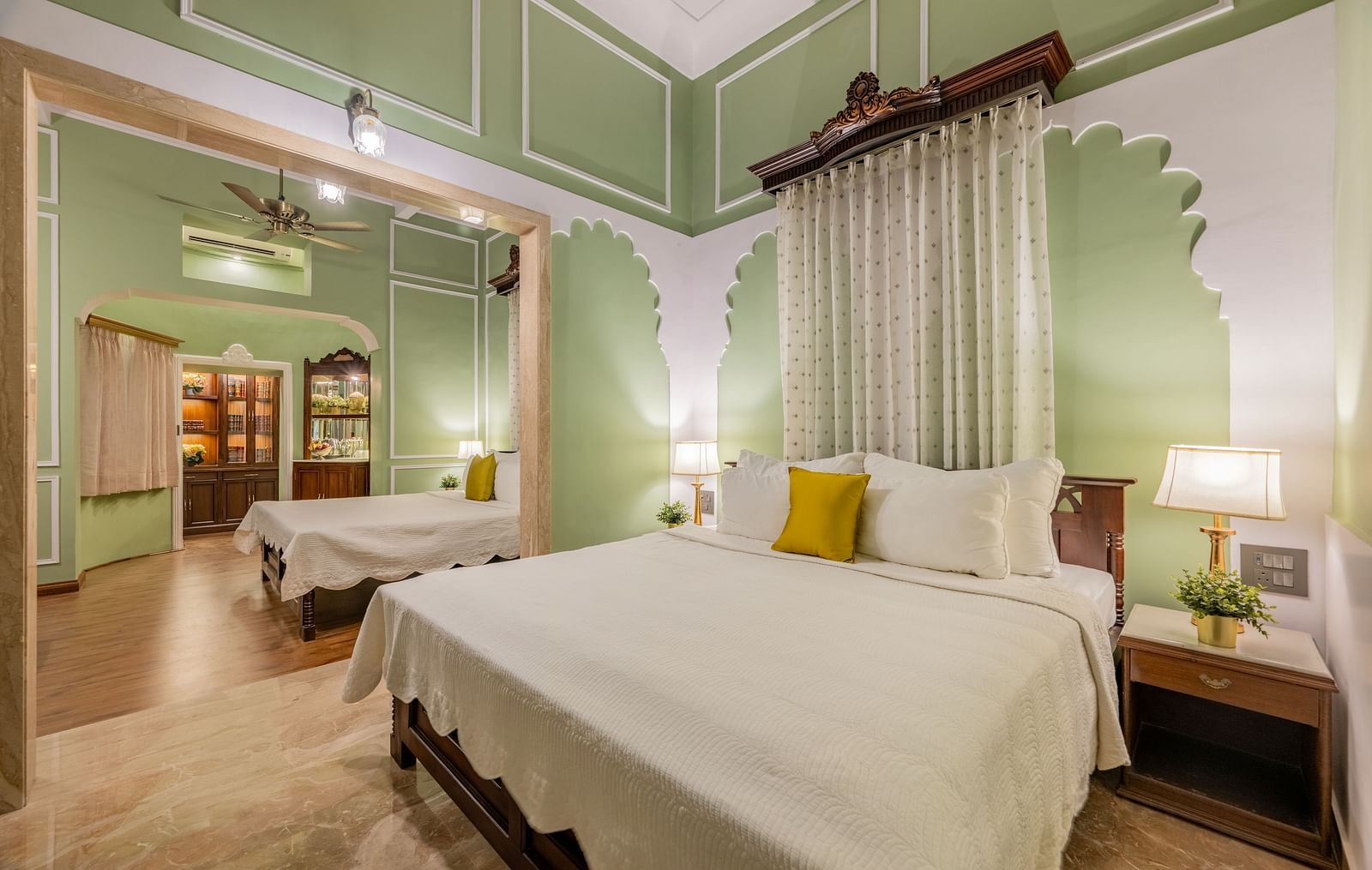 A serene suite at Dera Rawatsar, Jaipur, features a comfortable four-poster bed, an additional single bed in an adjacent area, and elegant wall paneling.