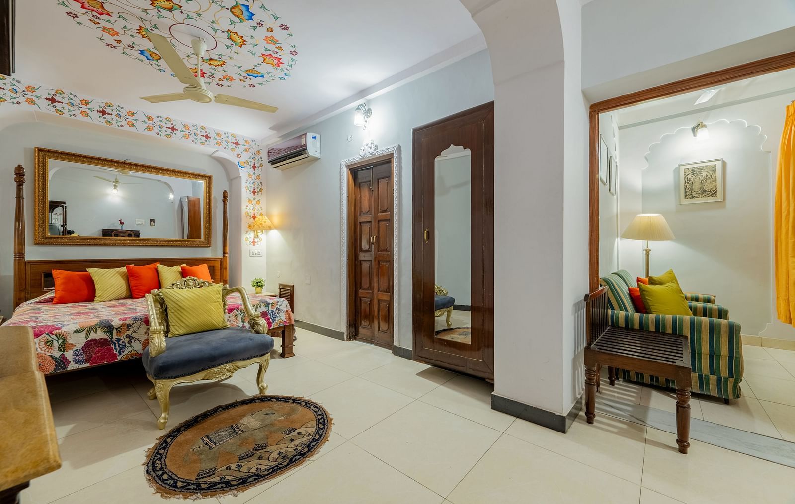 A room with a patterned ceiling, a large mirror, a bed with colorful cushions, a blue armchair, and a doorway leading to another room | Dera Rawatsar, Jaipur