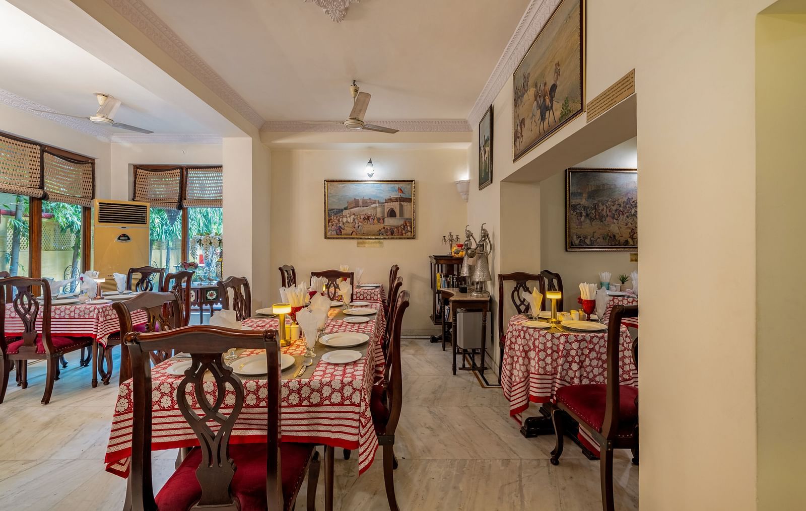 The dining area at Dera Rawatsar, Jaipur, features set tables with patterned cloths, wooden chairs, and artwork adorning the walls.