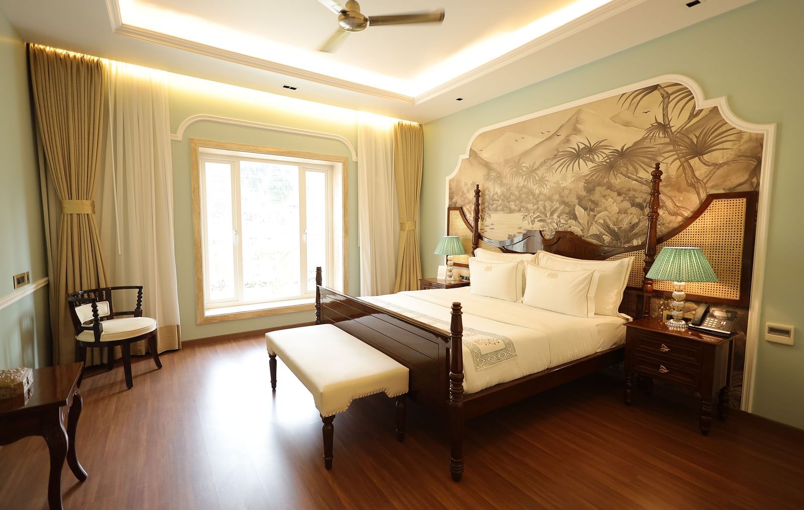 A bedroom featuring a four-poster bed, a large window with sheer curtains, a wall mural, and polished wooden floors, under a recessed ceiling with soft lighting | Dileep Kothi, Jaipur