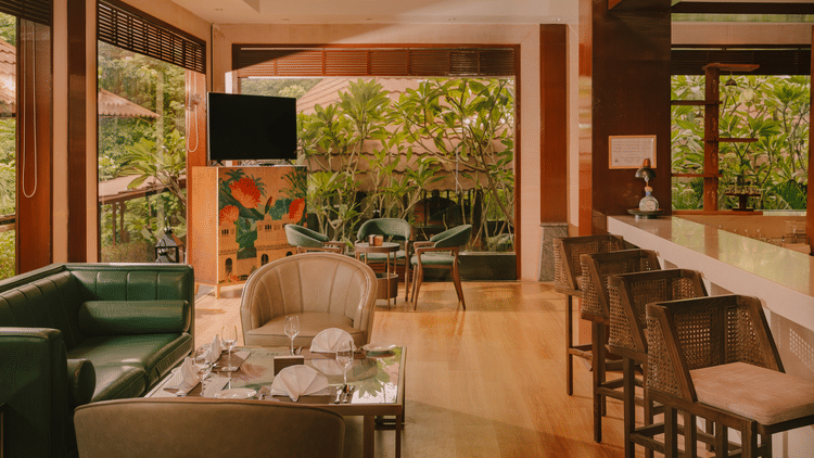 A well-lit lounge area at The Ananta Udaipur, featuring comfortable seating, a bar counter, and large windows overlooking the lush greenery.