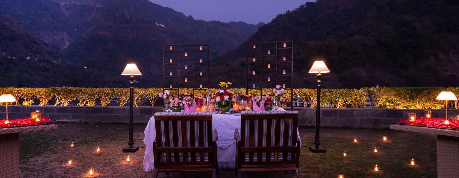 A dinner setup outdoors with a table adorned with candles and flowers,he backdrop is a beautiful mountain range at dusk.