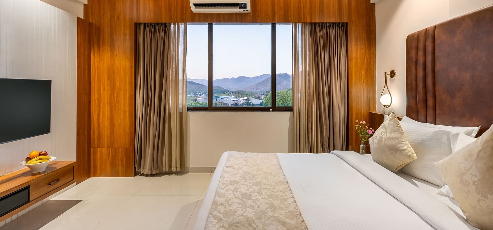 A comfortable king-size bed with soft pillows and a throw faces a window with curtains revealing a distant landscape - Lords Eco Inn Ambaji.