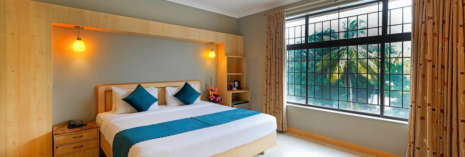Executive Room featuring a queen size bed and a night stand next to it as the sunlight from the expansive windows makes its way in at Cross Roads Inn, Bengaluru