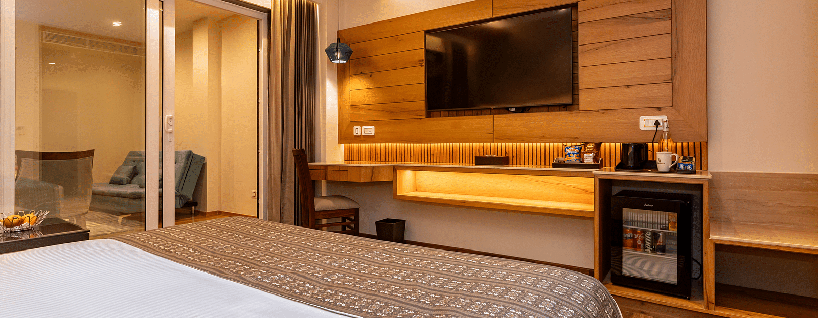 An image of a Family Room with bed, flat screen TV, work desk and chair, modern room amenities, and an expansive glass door leading to other areas of the room - The Belvedere Himalayan Retreat, McLeodganj