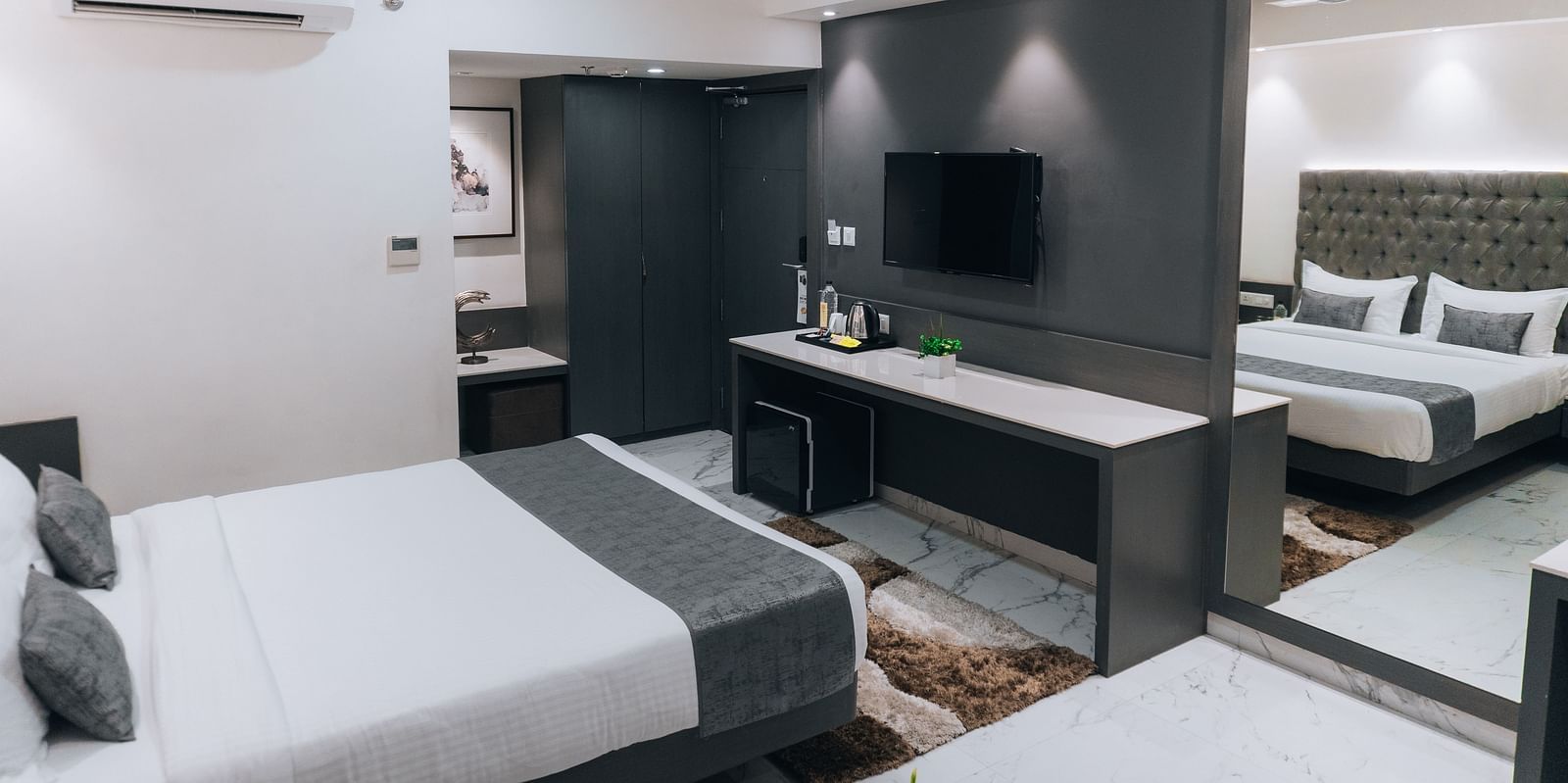 a bedroom with the bed facing a mounted television, with a workdesk beneath and a large mirror on the side - Jardin Hotels, Indore