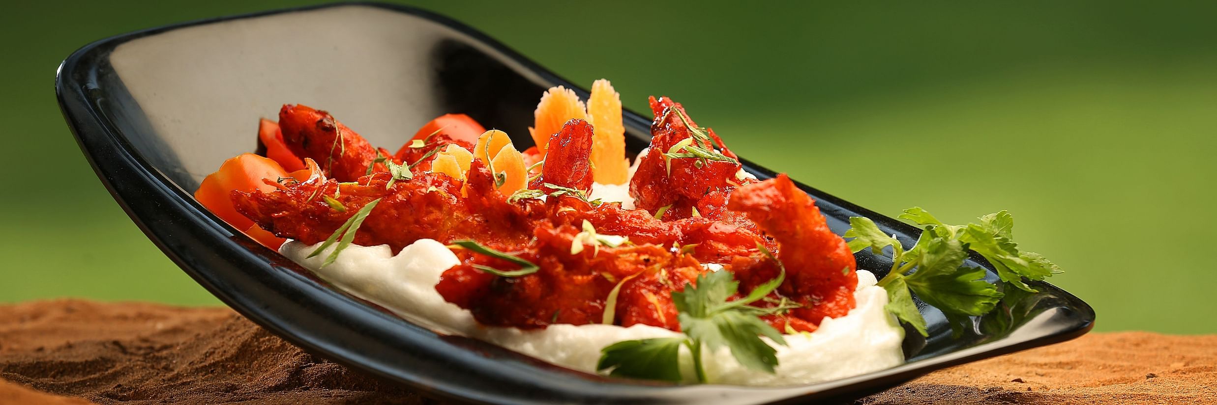 A black, angled plate on a wooden surface holds a vibrant dish of red, saucy prawns over a white base, garnished with herbs.