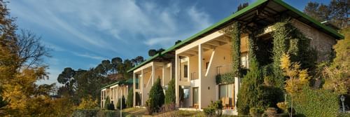 Facade of Suryavilas Luxury Resort and Spa in Solan with a paved driveway and green climbing plants on its exterior walls, under a blue sky with clouds
