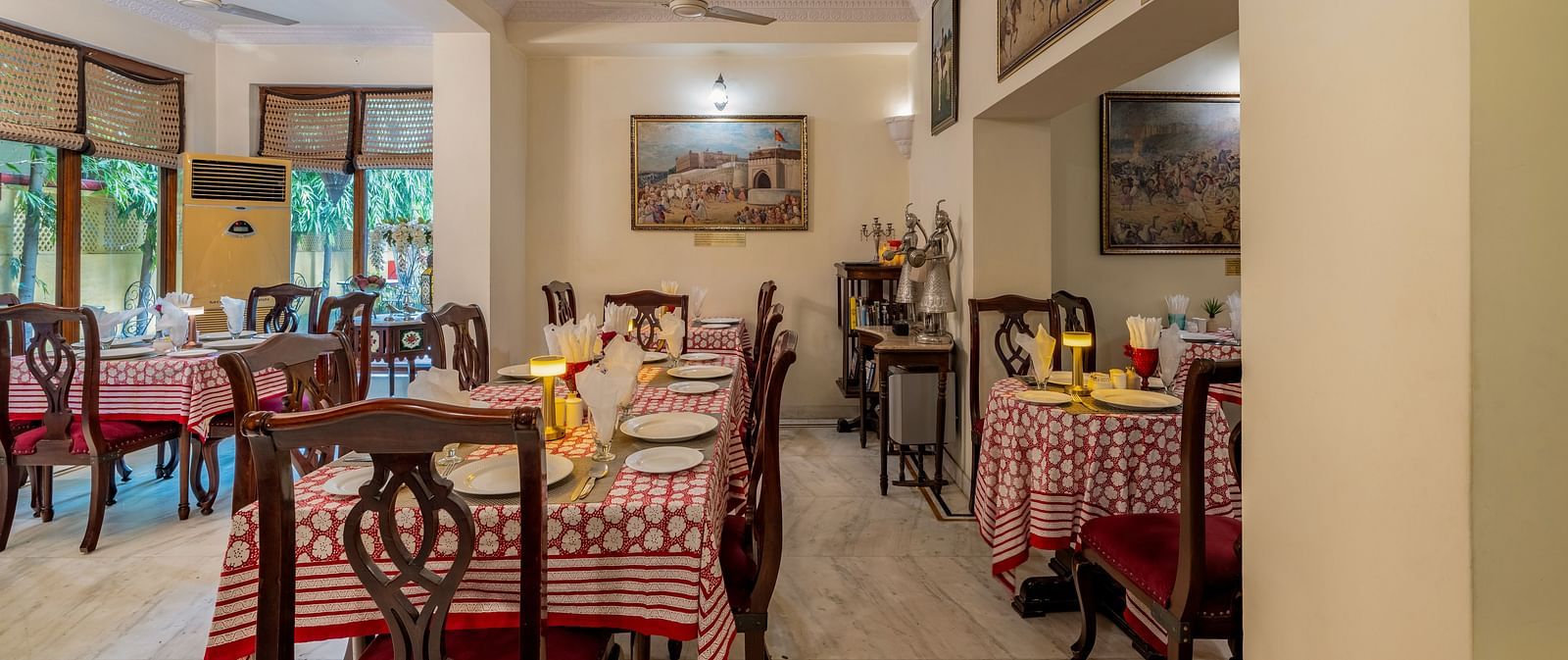 The dining area at Dera Rawatsar, Jaipur, features set tables with patterned cloths, wooden chairs, and artwork adorning the walls.