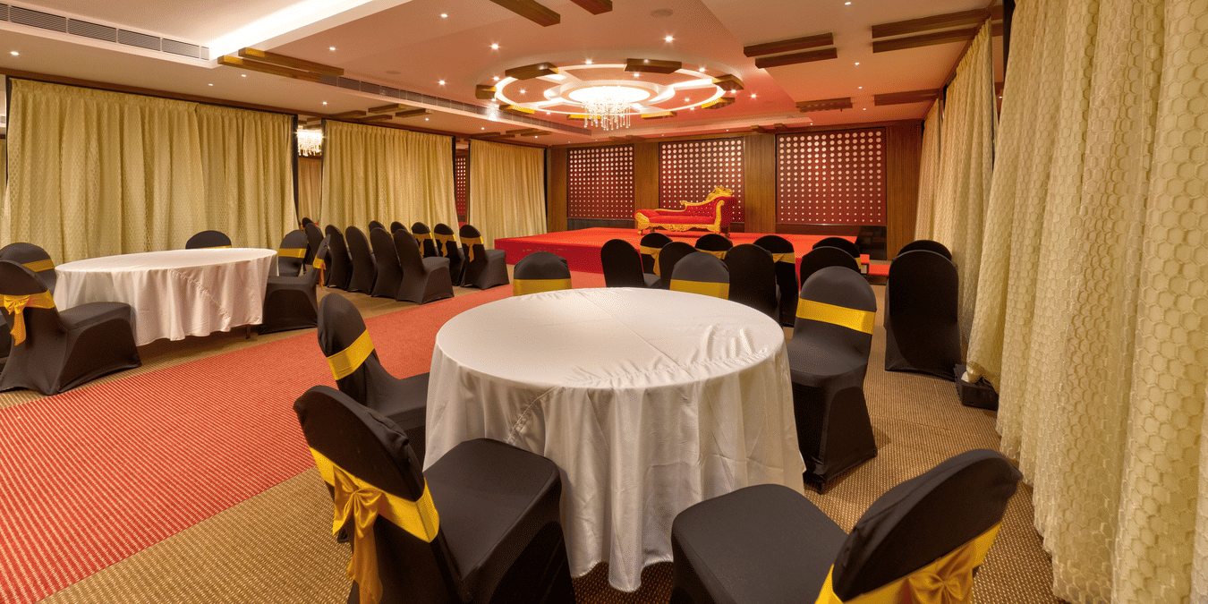 Round tables, black and gold chairs, a red carpet, and a stage setup inside the banquet hall of Icon Express By Bhagini, Hoodi.