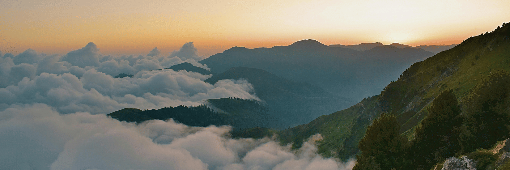 Sunset view from a mountain peak overlooking a sea of clouds and rolling green ridges.