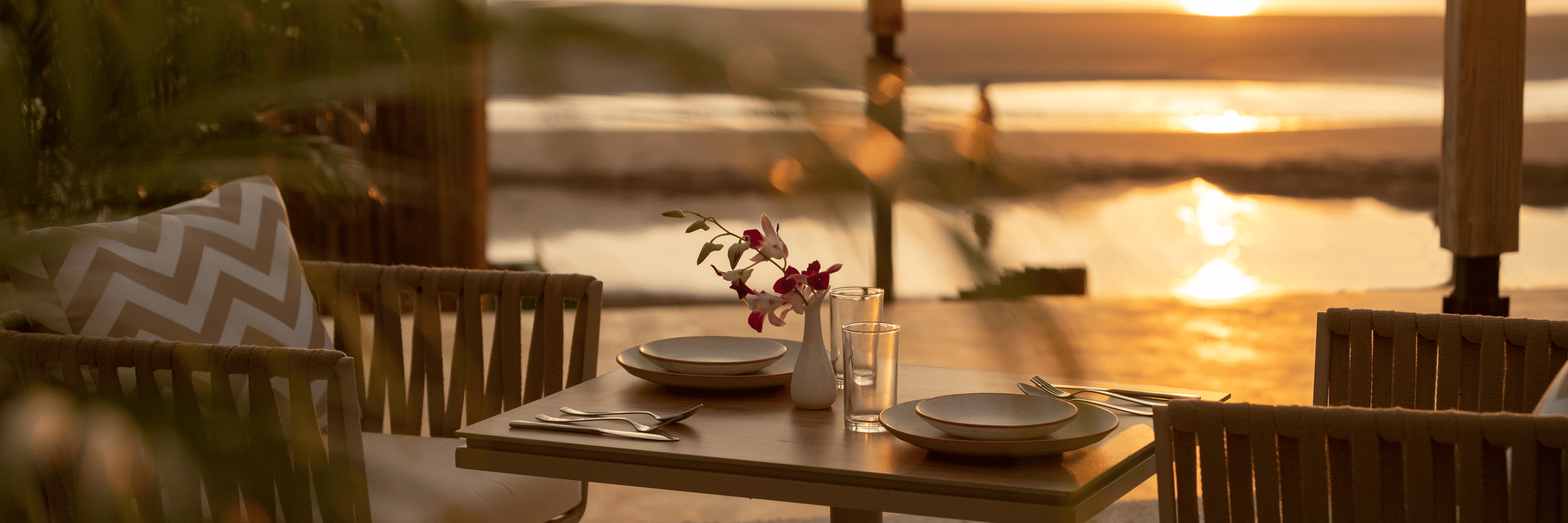 A close up shot of a table with two chairs at saz on the beach restaurant during sunset with sun rays reflecting on the sea waves in the background