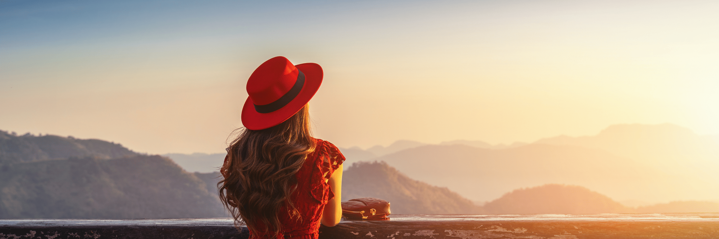 Person in a red outfit and hat sits on a stool at a scenic overlook, gazing at layered mountains during sunrise or sunset, with a drink placed on the counter in front.