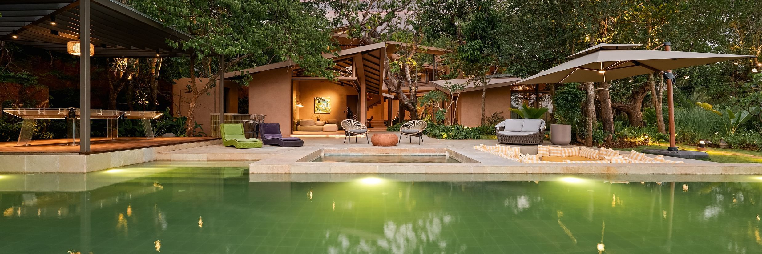  A beautiful evening view of Villa Moira by V Escapes, showcasing the illuminated green pool and the villa's warm lighting under a colourful twilight sky.