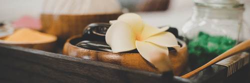 Spa essentials with herbal ingredients, a flower, and massage tools arranged in a wooden tray for wellness therapy.