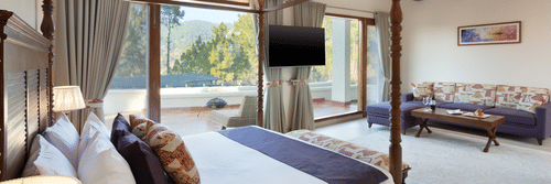 Spacious luxury resort room with a four-poster bed, large windows offering scenic outdoor views, a wall-mounted TV, and a comfortable sitting area with natural light streaming at Suryavilas Luxury Resort and Spa
