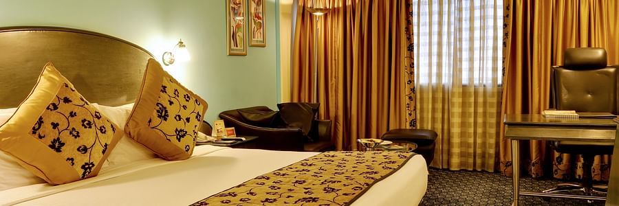 An image of a bedroom with double size bed, cushions placed on top, there is a chair and a work desk at one corner, and at another corner there is a sofa for seating