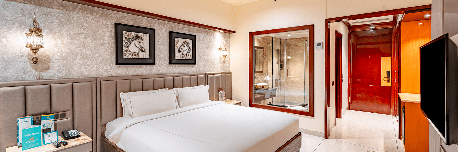 A spacious bed with two photo frames above, a bedside table with essentials, and a wall mounted TV opposite at  Crescent Spa & Resort, Indore