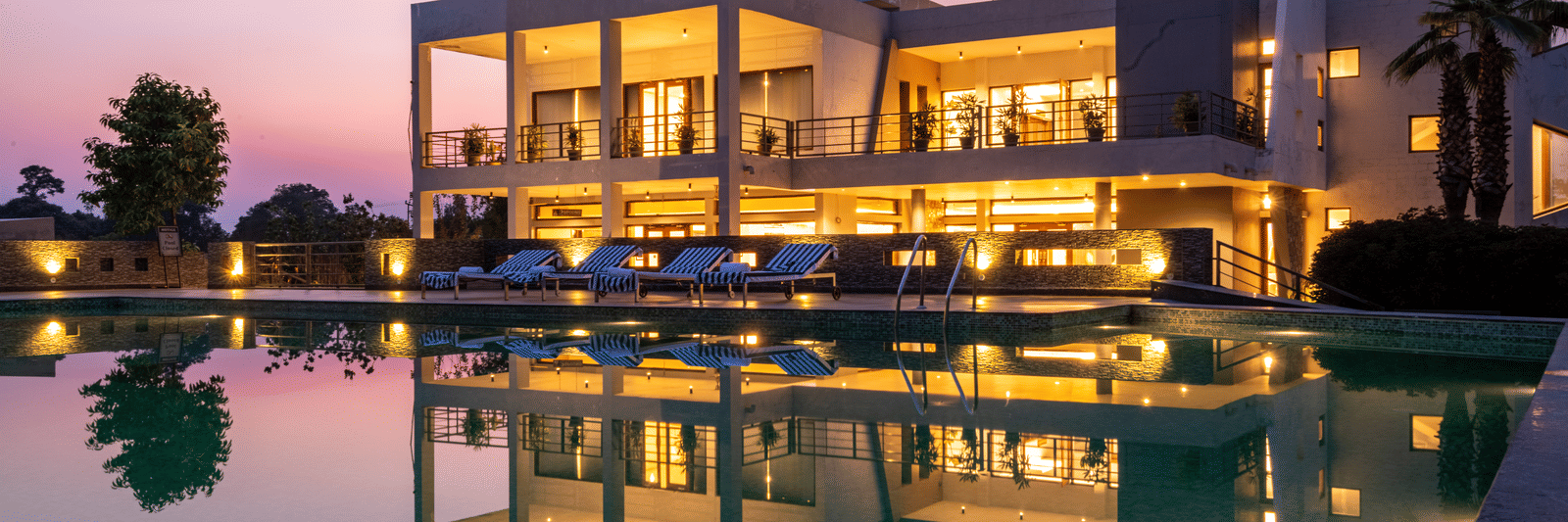 Facade view of DLS Forest Vines Luxury Resort in Jim Corbett, with warm light glowing from its windows and doors, reflected on the shimmering swimming pool beneath a clear sky.