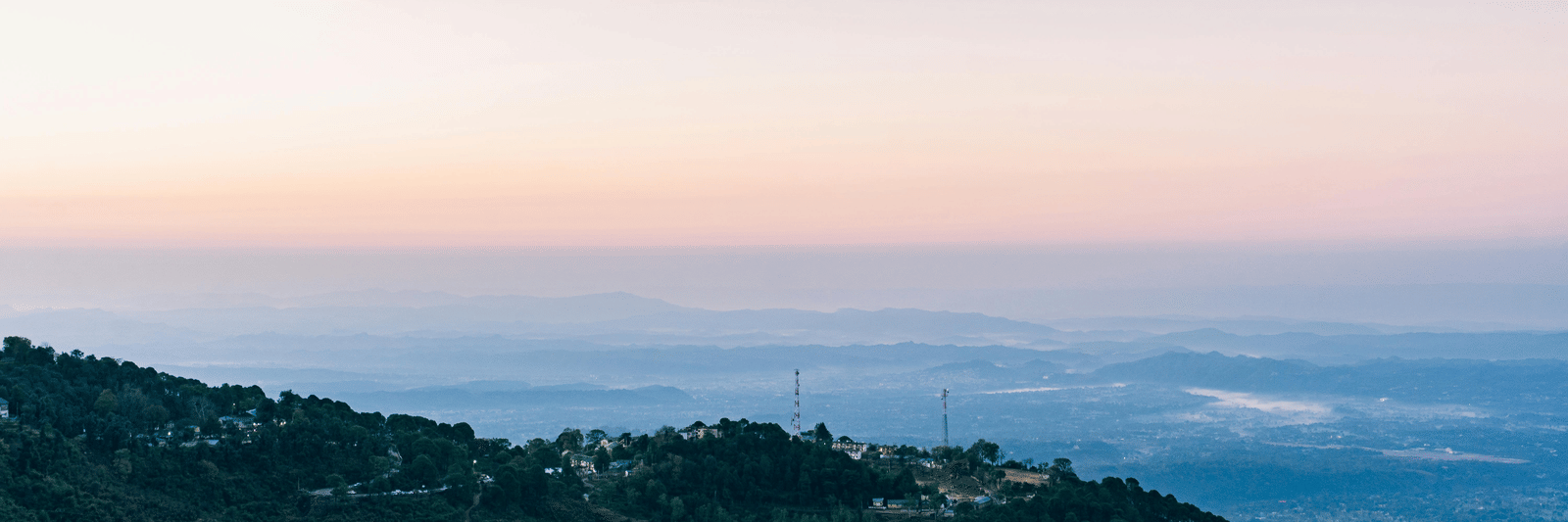 Scenic view of a hillside town with forested slopes, overlooking a vast valley and distant mountains under a pastel sky.