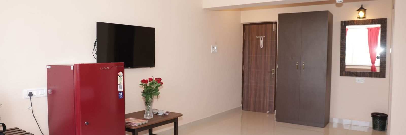 A spacious room at Nazri The Verda Hotel, Arambol, displaying a table with flowers, a TV mounted on the wall, and a red fridge.