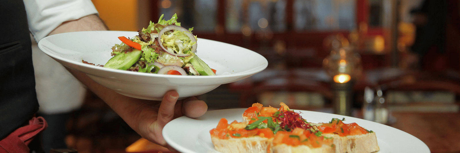 A person in a restaurant holding two plates with delicious dishes in it that are well plated.