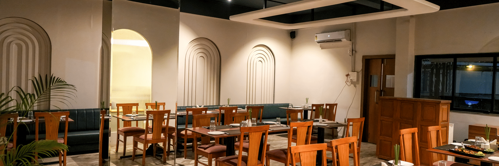  An interior view of the bright dining hall with white walls, wooden chairs, and recessed archways at Pravasa Hotel, Kolhapur.