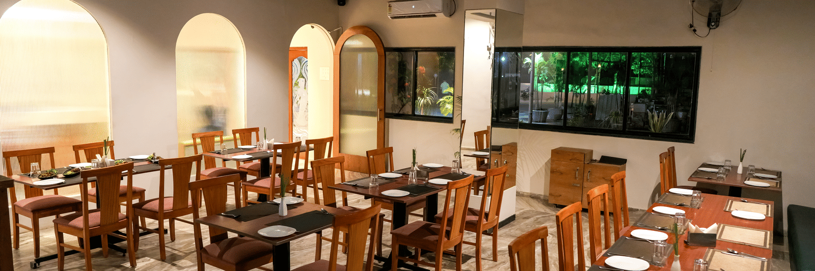 A well-appointed indoor dining room with wooden tables and chairs, white walls, and elegant archways at Pravasa Hotel, Kolhapur.