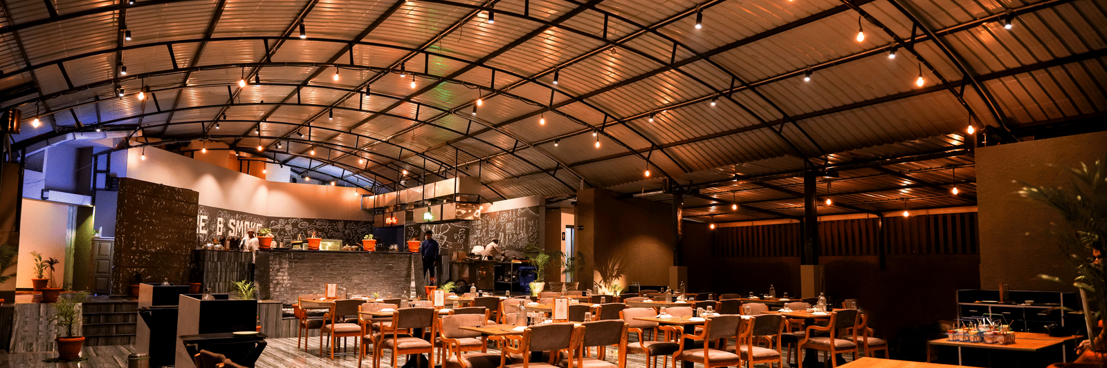  A wide view of the spacious dining hall with wooden tables and chairs under a curved, industrial-style roof at Pravasa Hotel, Kolhapur.