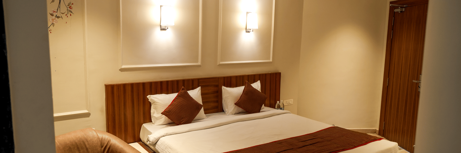  A modern guest bedroom at Pravasa Hotel, Kolhapur, featuring a double bed, subtle wall panel moulding, and a tan armchair. 
