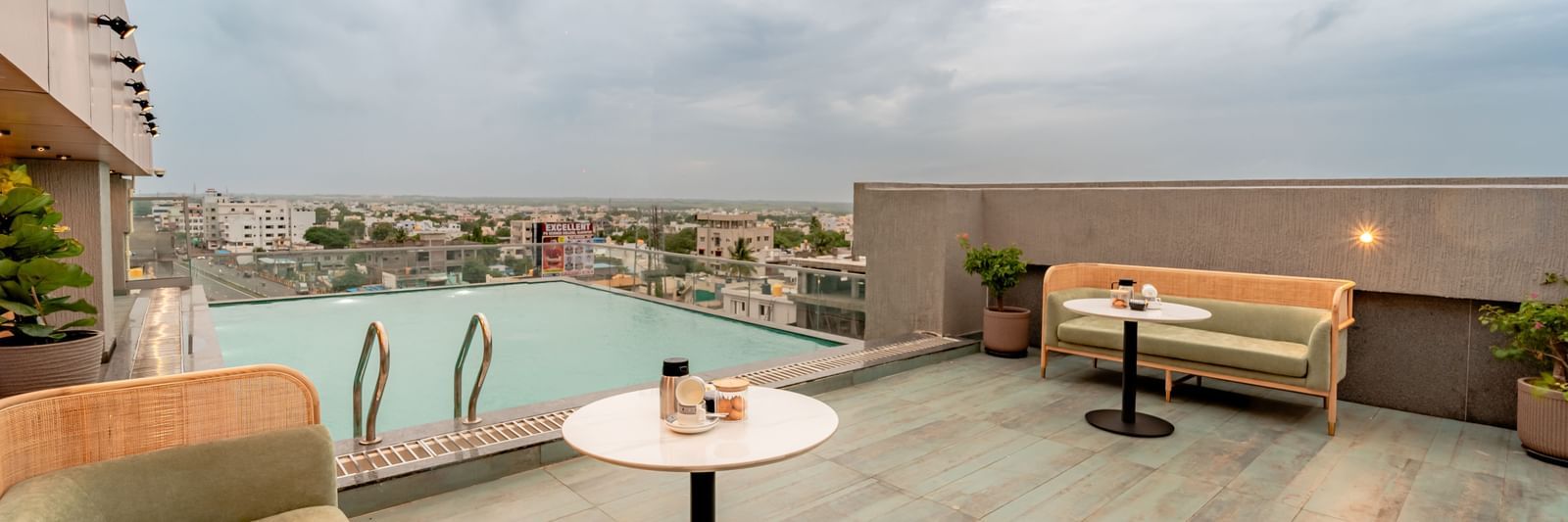 The rooftop swimming pool with clear water, surrounded by a tiled deck and some modern outdoor seating, offering a panoramic view of a city under a cloudy sky | Pravasa Hotel Vijayapura