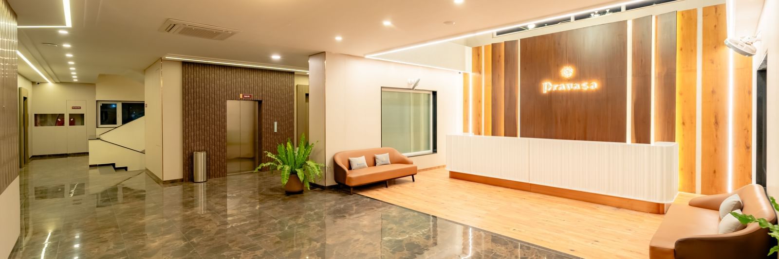 A well-lit waiting lounge with light-colored sofas and a large potted plant | Pravasa Hotel, Vijayapura