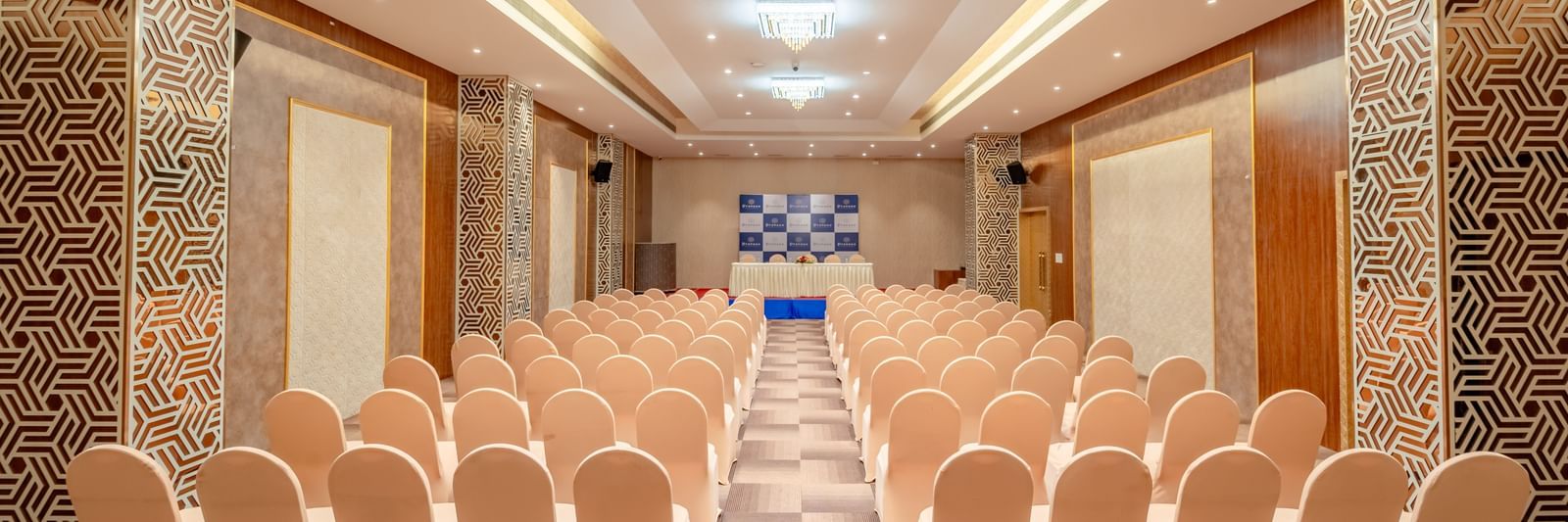 An empty event space features multiple rows of chairs and a prominent stage area | Meetings & Events | Pravasa Hotel, Vijayapura