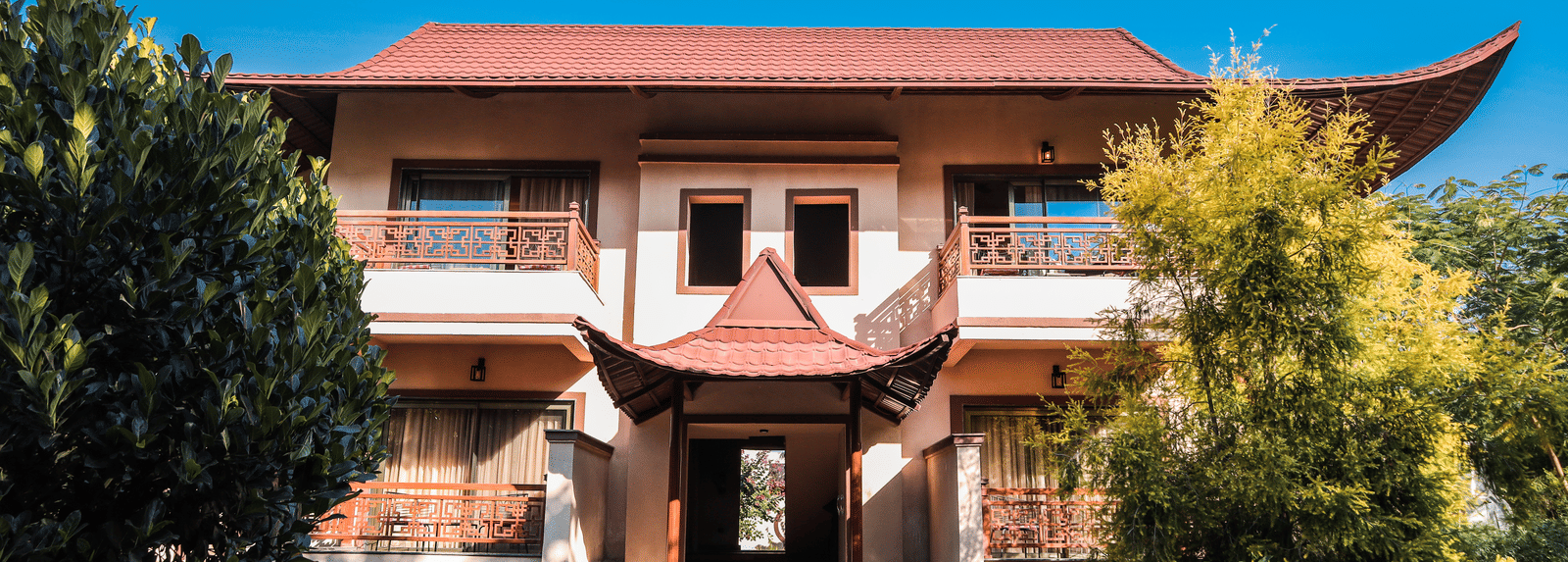 Two-storey building with Asian-style red tiled roof, decorative balconies, and a central entrance, surrounded by trees, plants, and a garden under a clear blue sky - Ananta Spa & Resort, Jaipur