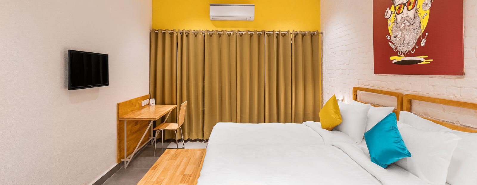 A side-angle interior image of the deluxe room at Bedzzz Varanasi showcases a spacious layout featuring a large bed, a window with elegant curtains, a TV, a decorative wall poster, ample lighting, and a cupboard for storage.