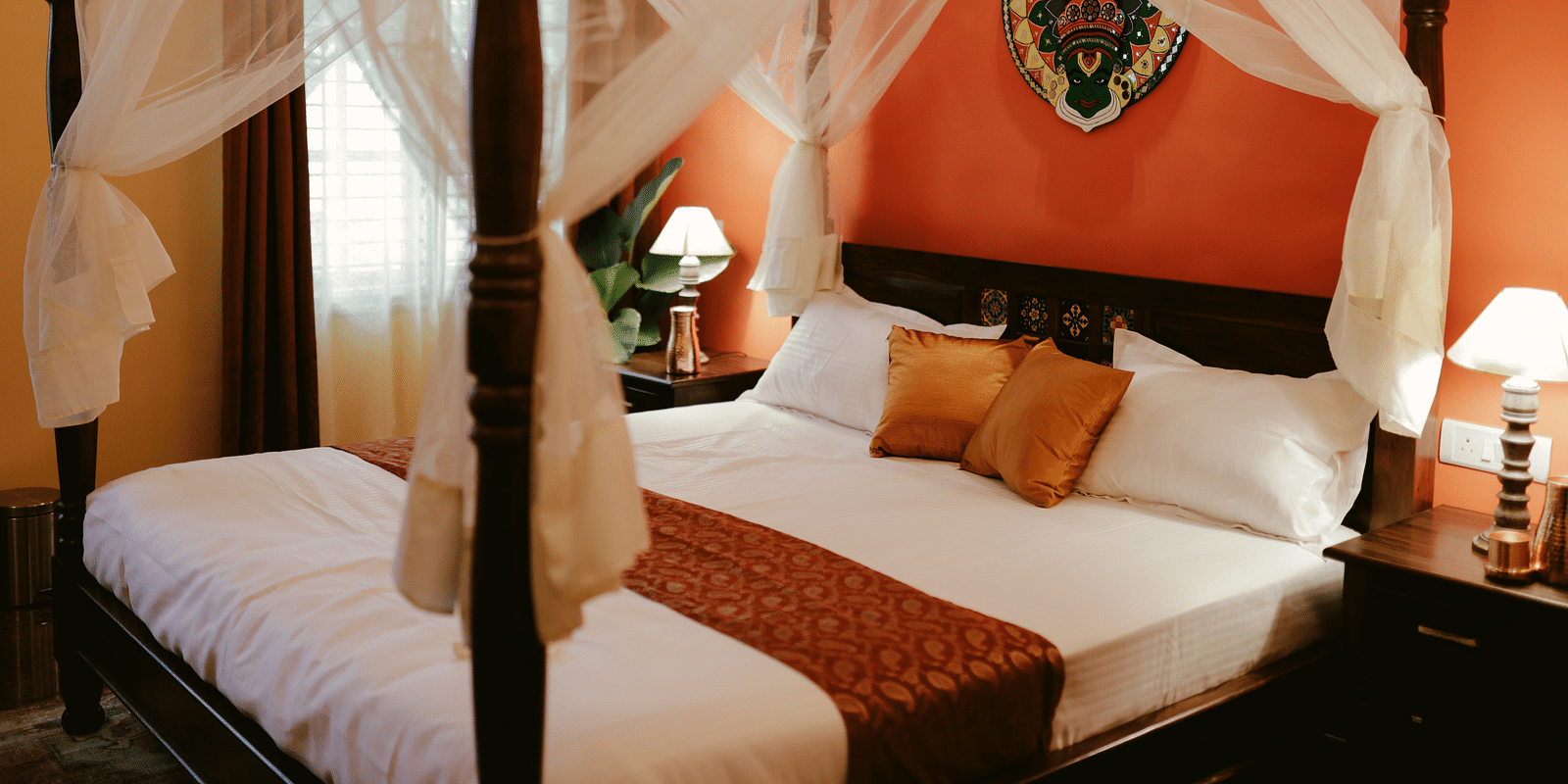 A four-poster bed with white drapery and a decorative headboard in a room with an orange accent wall | Adhyaya Heritage Reverbed