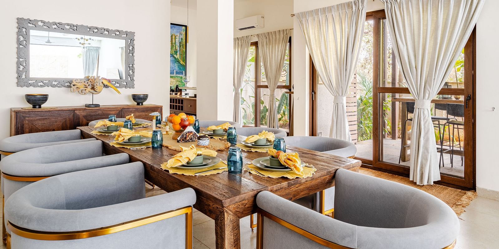 Dining space at Lindsay Manor with a large wooden table, cushioned chairs, hanging lights, and large windows with curtains.