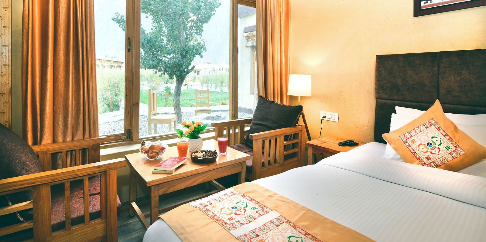 Standard Cottage with a king size bed, a window with orange curtains, a small table and chairs, and a painting on the wall at White Sands Resort, Ladakh.