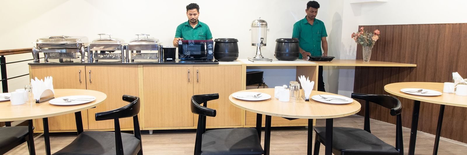 A breakfast buffet area with staff behind the counter, food warmers, and tables with black chairs - Sanctum Suites Richmond Road, Bangalore