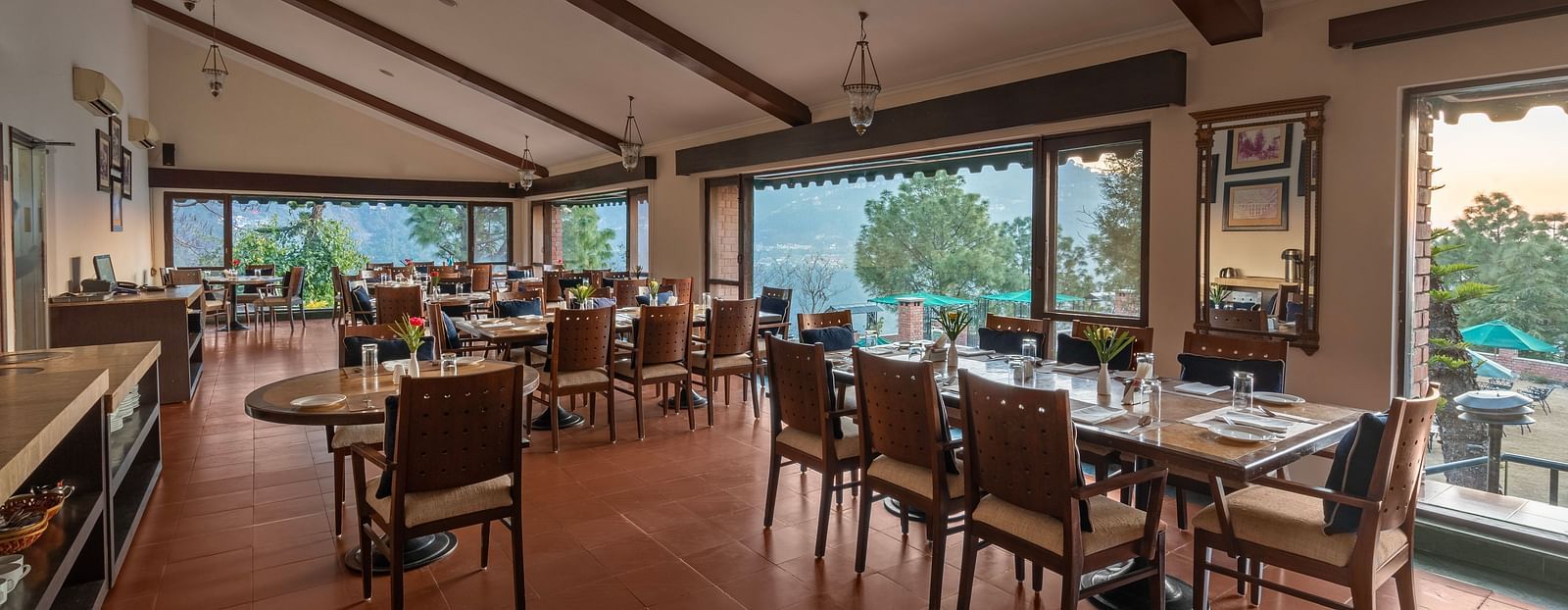 A restaurant with glass windows offering a beautiful mountain view, featuring dining tables arranged with chairs, a mirror on the wall, and stylish pendant lights overhead -  Baikunth resort, kasauli 