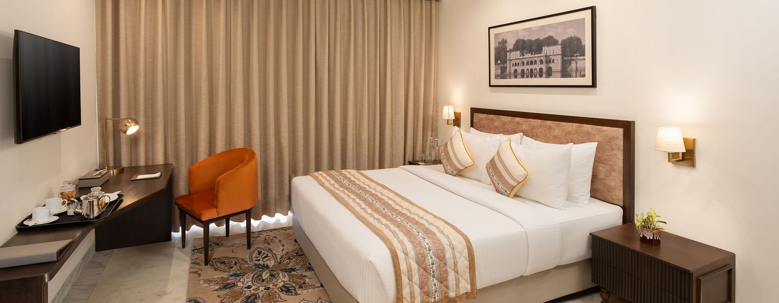 An image of a bedroom with bedside table, photo frame hanged on the wall, work desk with sofa chair, and a flat TV screen - Brij Eternity, Vrindavan