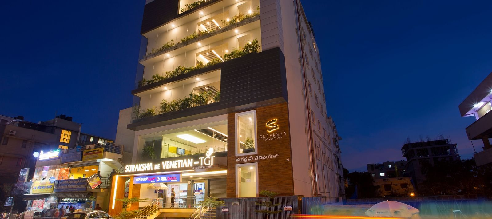 An exterior view of a modern multi-story building illuminated at dusk with signage and street activity - De Venetian by TGI, Brookefield