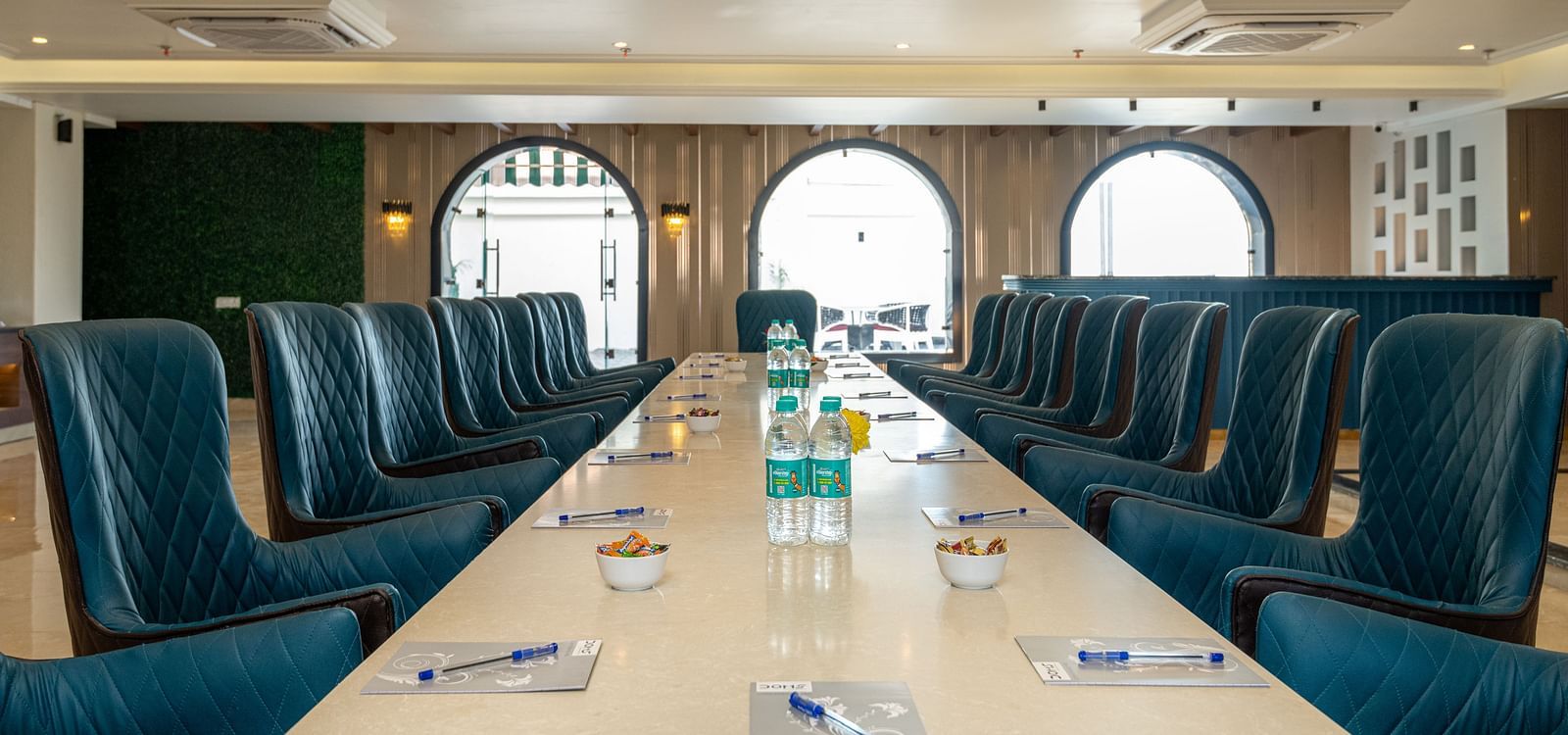 chairs arranged with a large table with notepads and pens along with water bottles on it as the daylight makes its way through the windows in the background