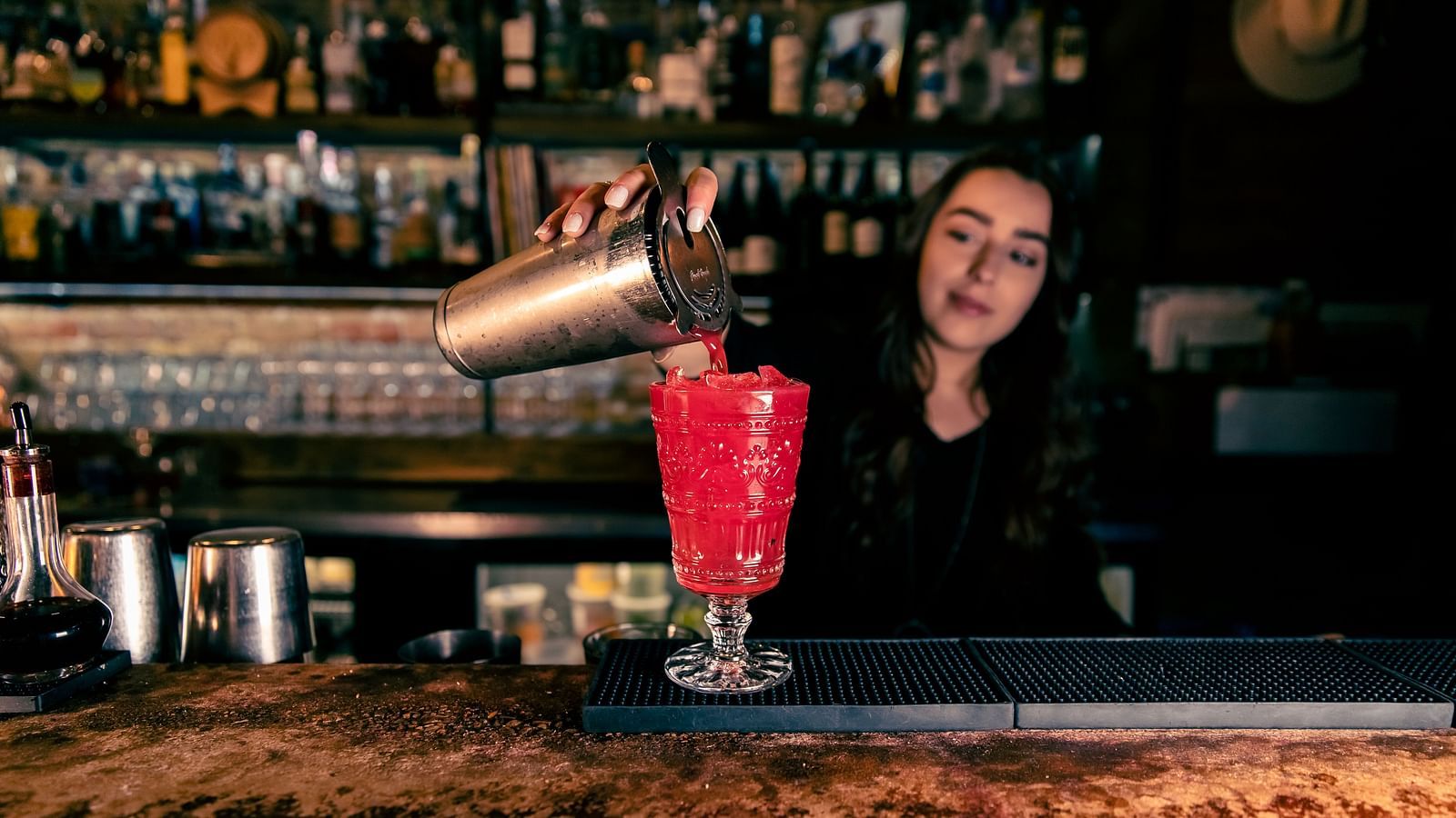 A bartender pours a drink into a glass at a bar, with shelves of liquor bottles in the background.- The Grand Hotel & Ballroom, McKinney